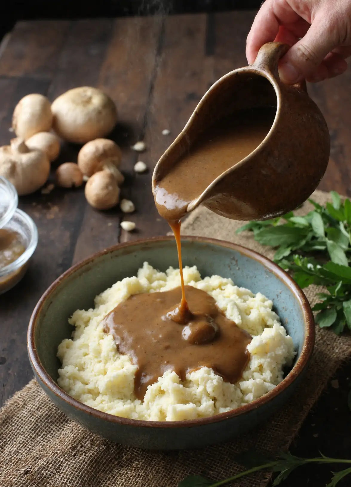 Mashed potatoes with gravy being poured from a small pitcher on a rustic wooden table.