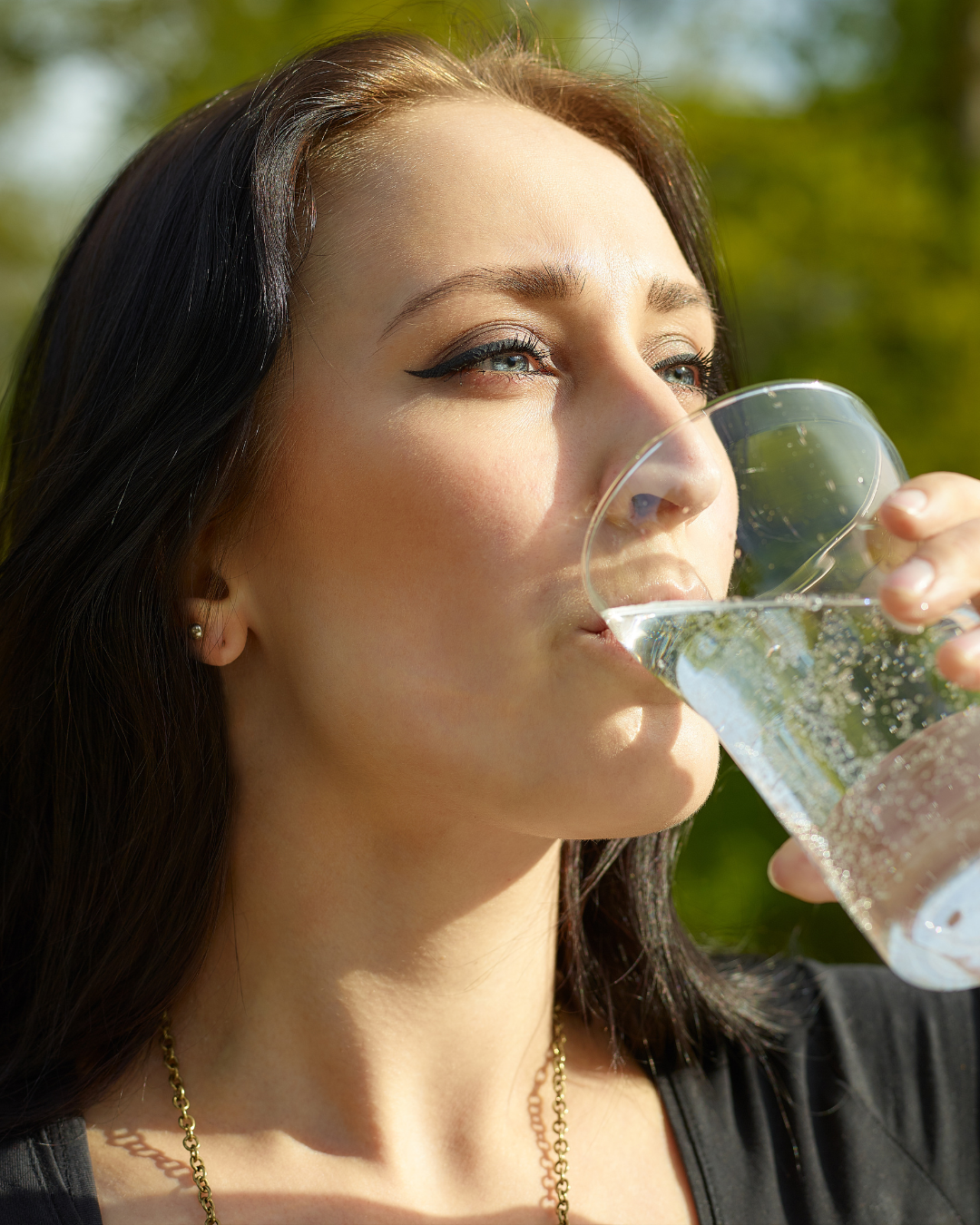Woman drinking from a glass outdoors with greenery in the background