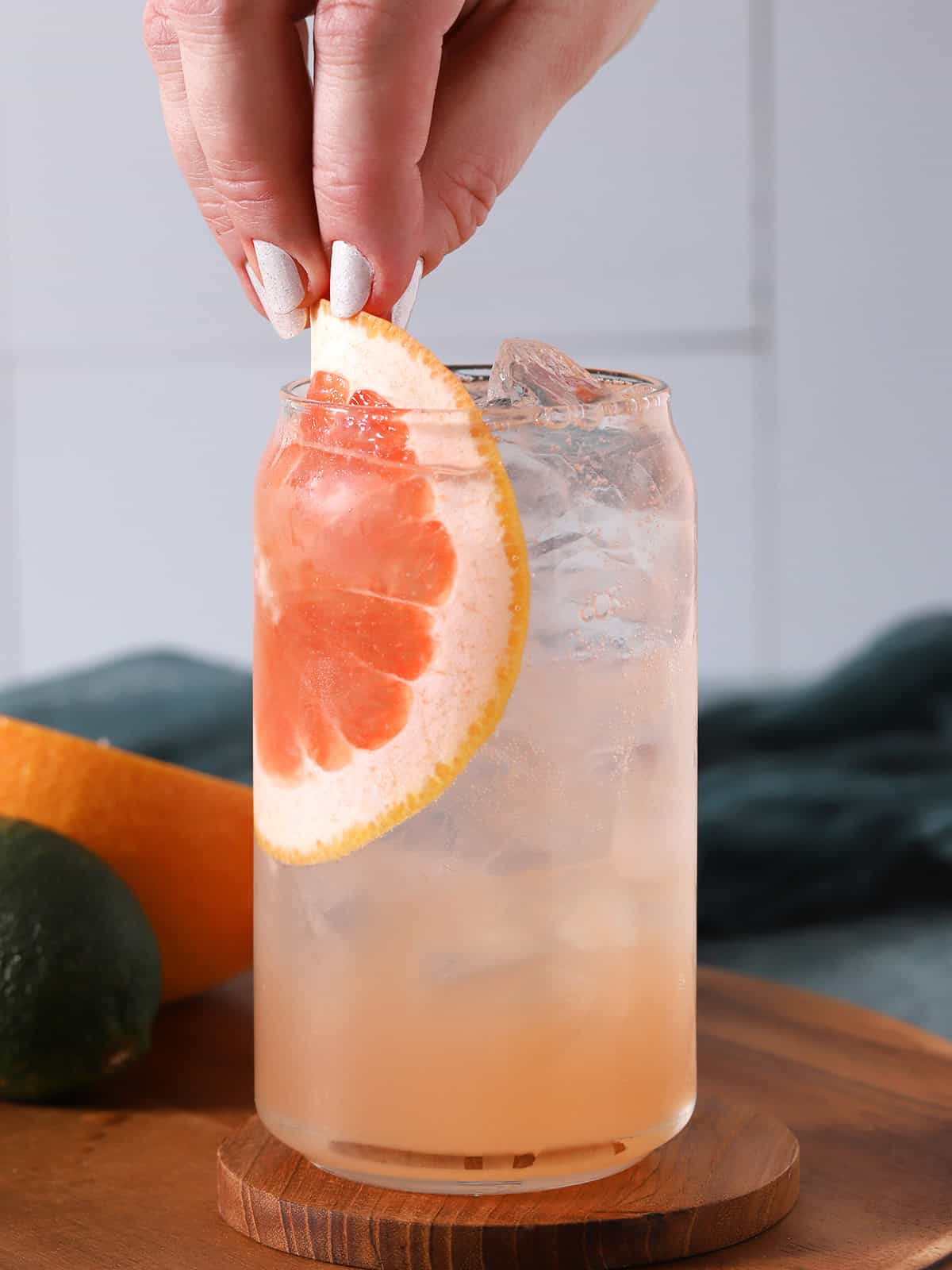 Cocktail with a grapefruit slice on a wooden coaster, with a hand placing the slice.