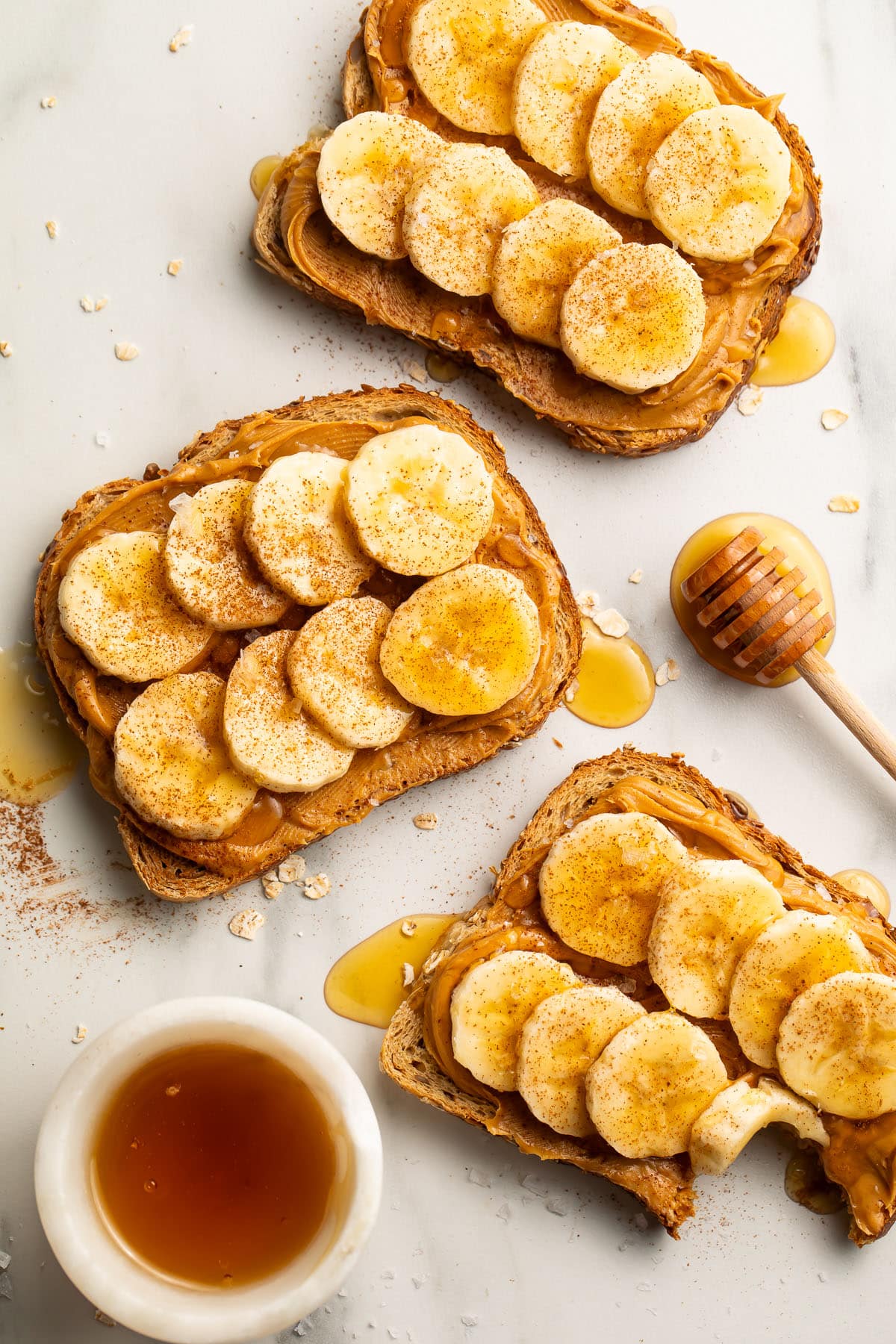 Three slices of toast with banana slices, peanut butter and honey on a light surface.