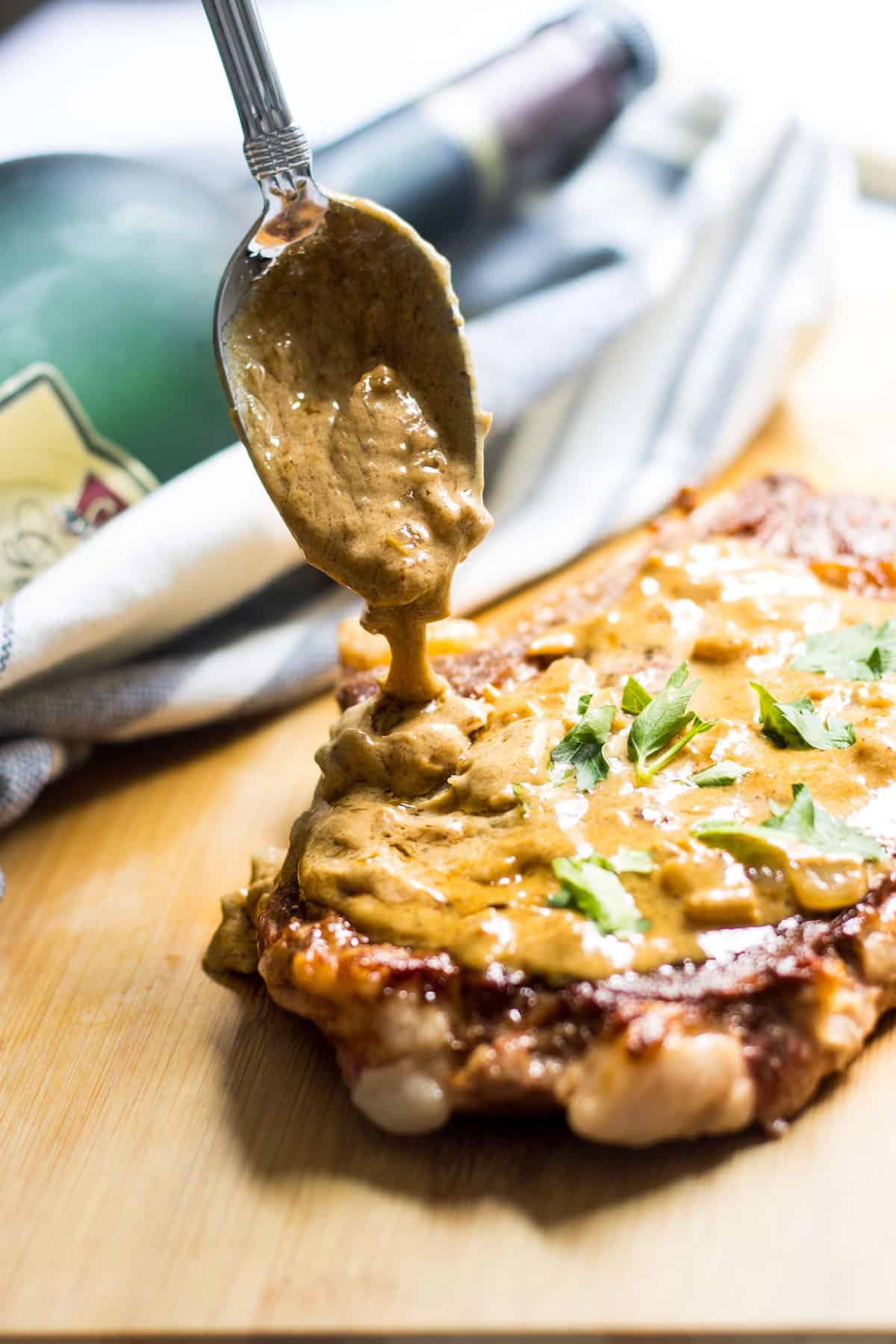 Spoon drizzling old fashioned dijon mustard over a steak on a wooden board with a bottle in the background.